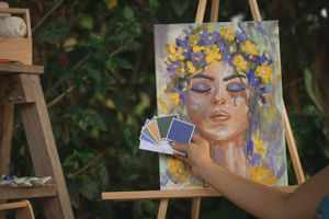 Hand holding color swatches against vibrant portrait painting with yellow and purple flower crown.