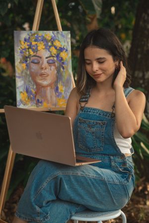 Artist in denim overalls works on laptop while sitting outdoors with their artwork displayed on an easel.