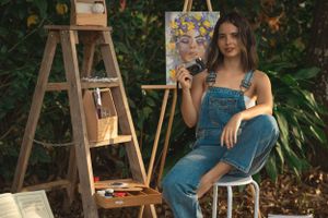 Artist in denim overalls sits outdoors with painting supplies and wooden easel.