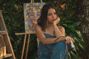 An artist sits thoughtfully next to an easel in an outdoor garden setting.