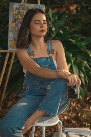 Artistic portrait in a garden setting with painting and camera, wearing casual denim overalls.