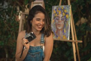 An artist in denim overalls poses with a vintage camera in front of their floral painting outdoors.
