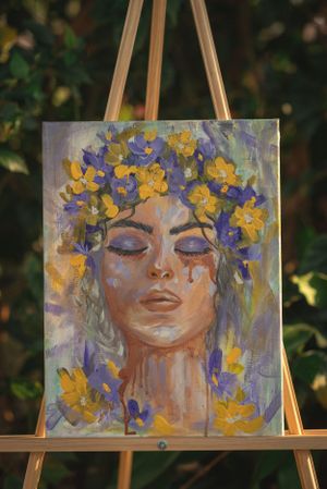 Painting of a serene face adorned with yellow and purple flowers on an easel outdoors.