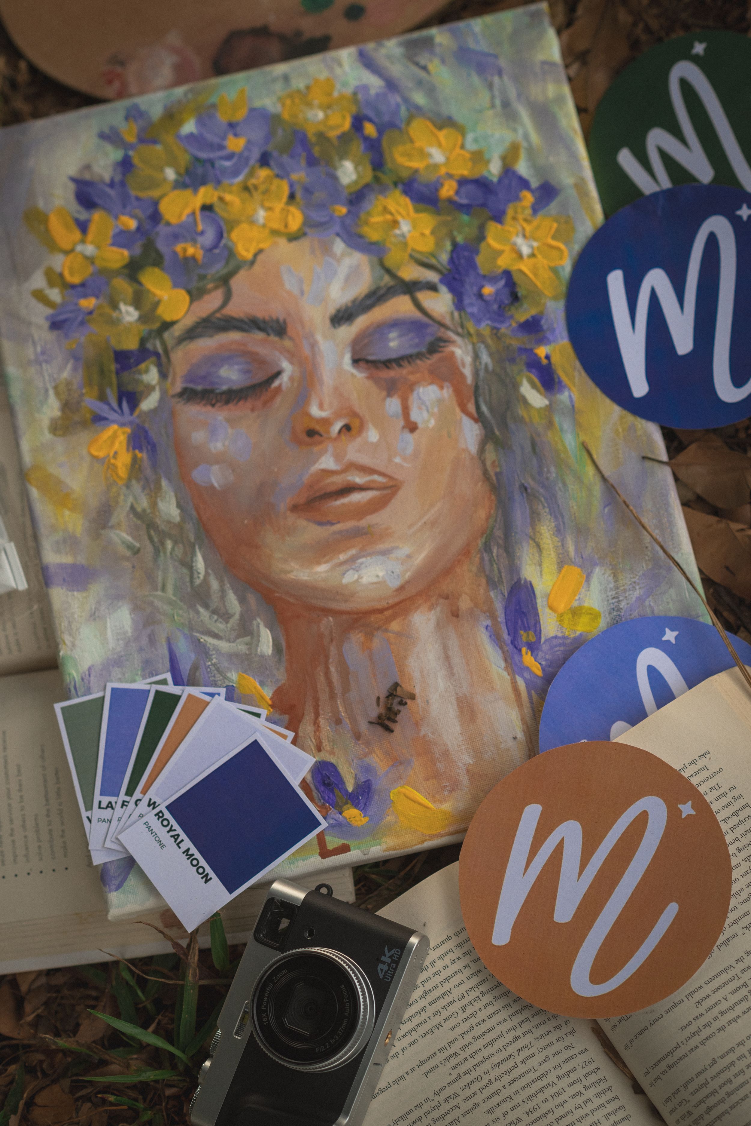 Artistic painting of a serene figure adorned with yellow and purple flowers resting on books with color swatches and a camera.