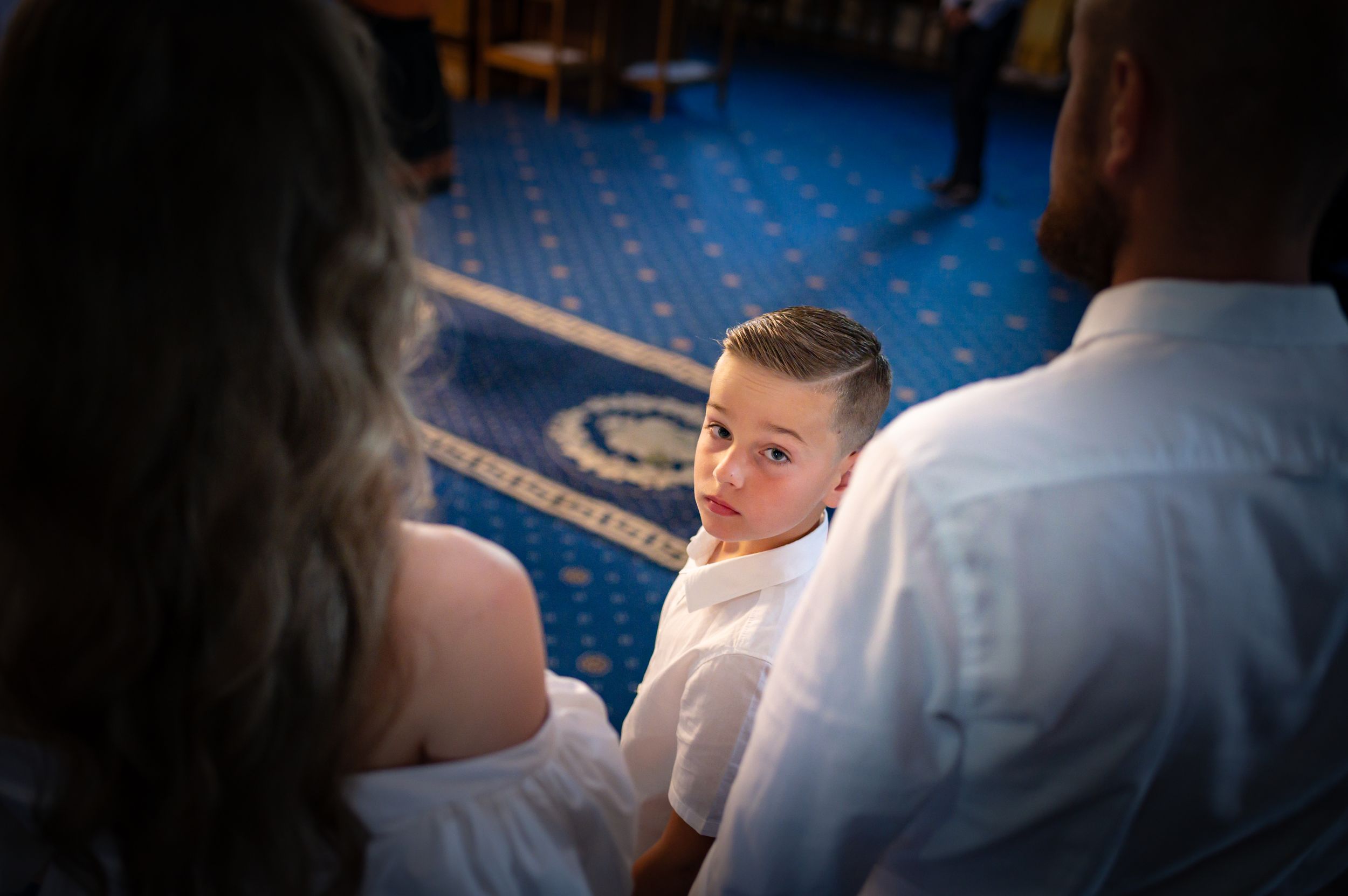 Young child looking back at parents during a formal ceremony on blue carpeted floor.