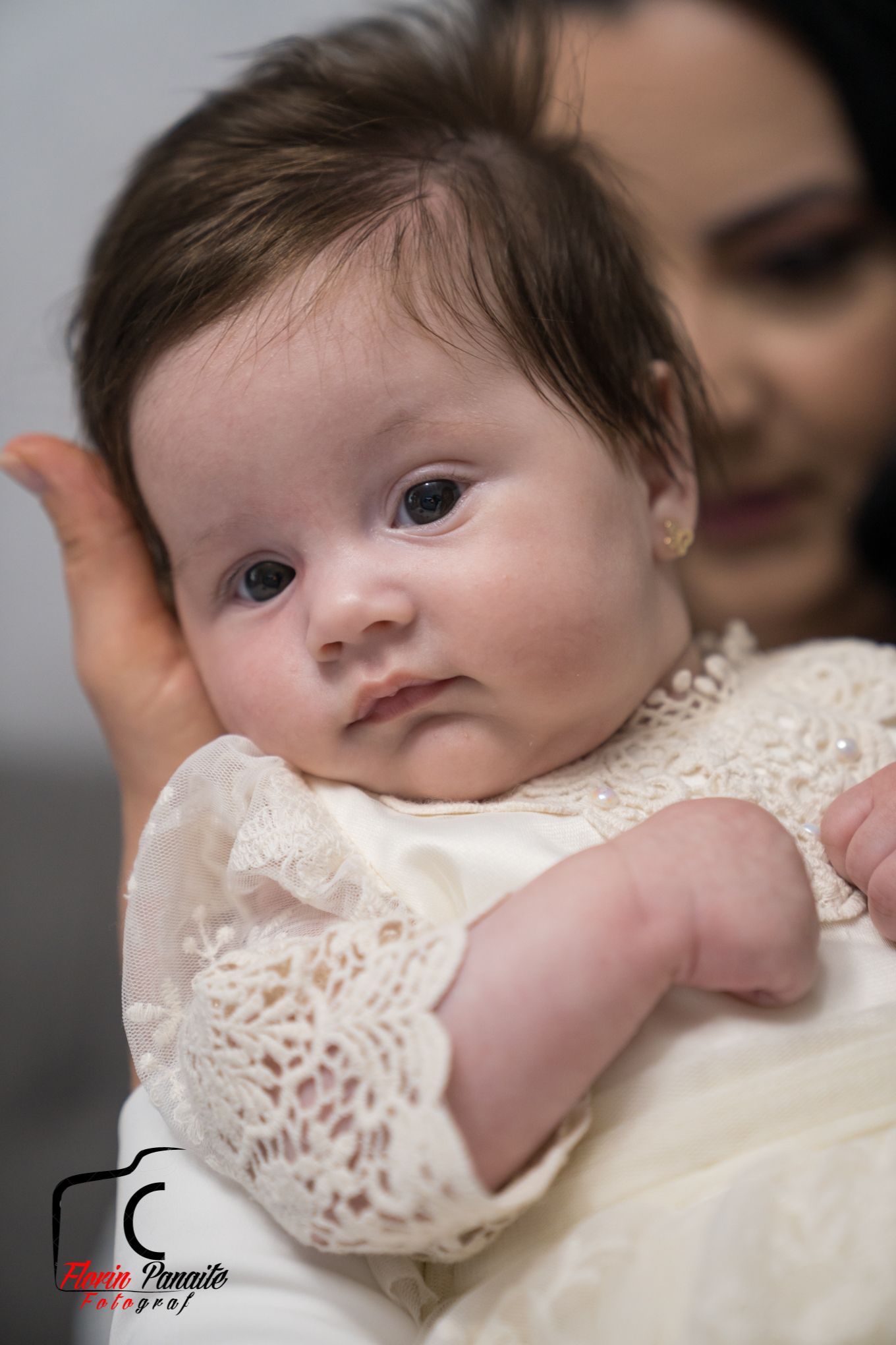 Baby wearing white lace christening gown looking directly at camera.
