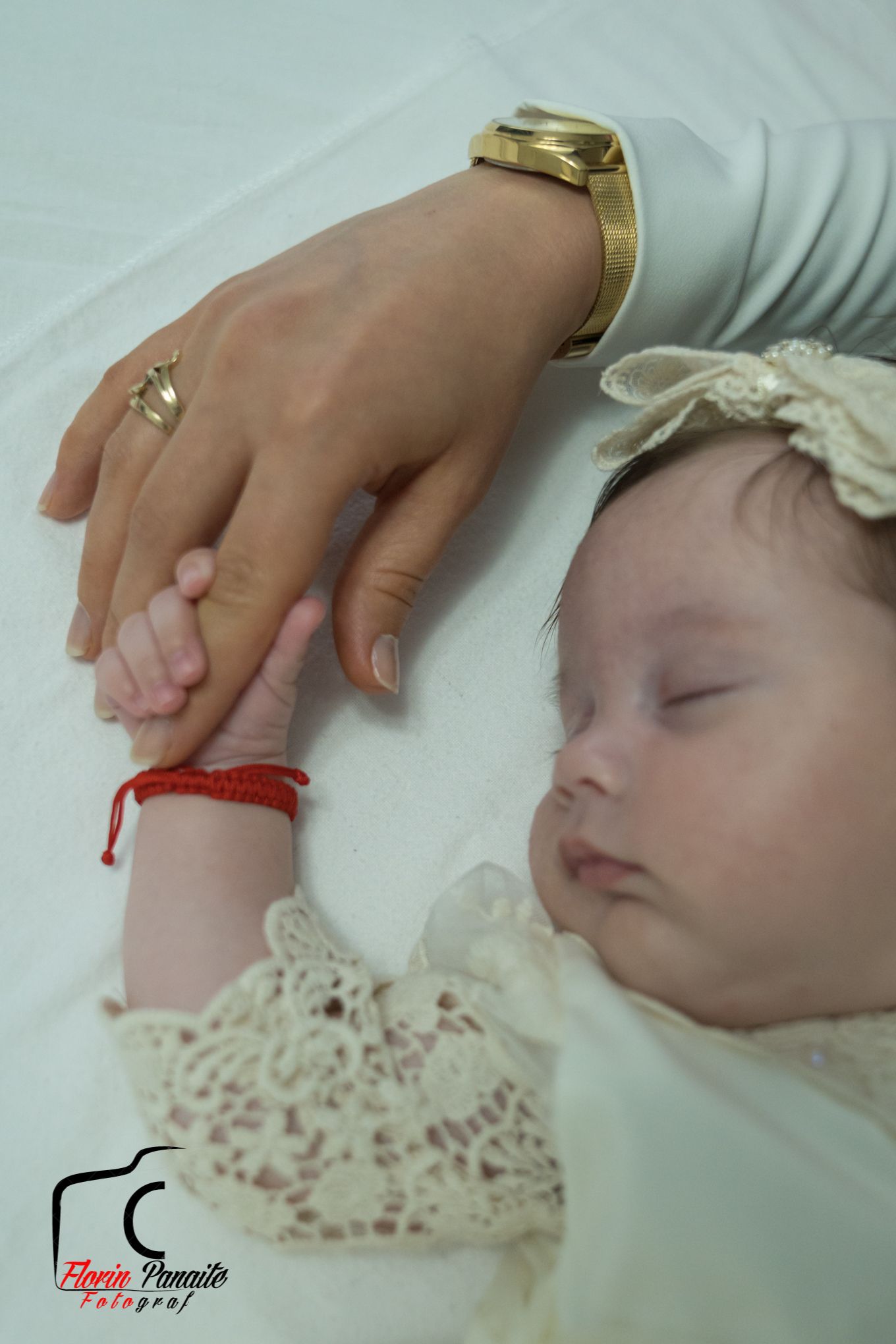 Sleeping baby in lace dress with adult hand wearing gold bracelet touching their arm.