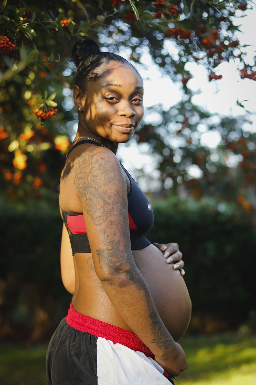 A maternity photoshoot outdoors with a person in a white and red dress posing against autumn foliage at sunset.