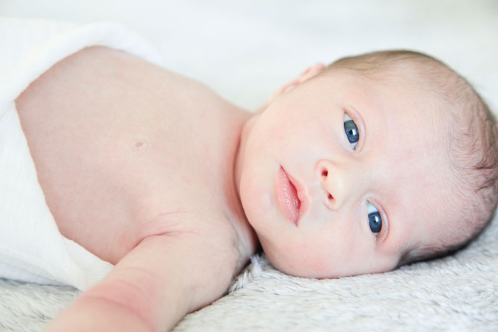 Baby with striking blue eyes lying on white bedding in a series of gentle portraits.