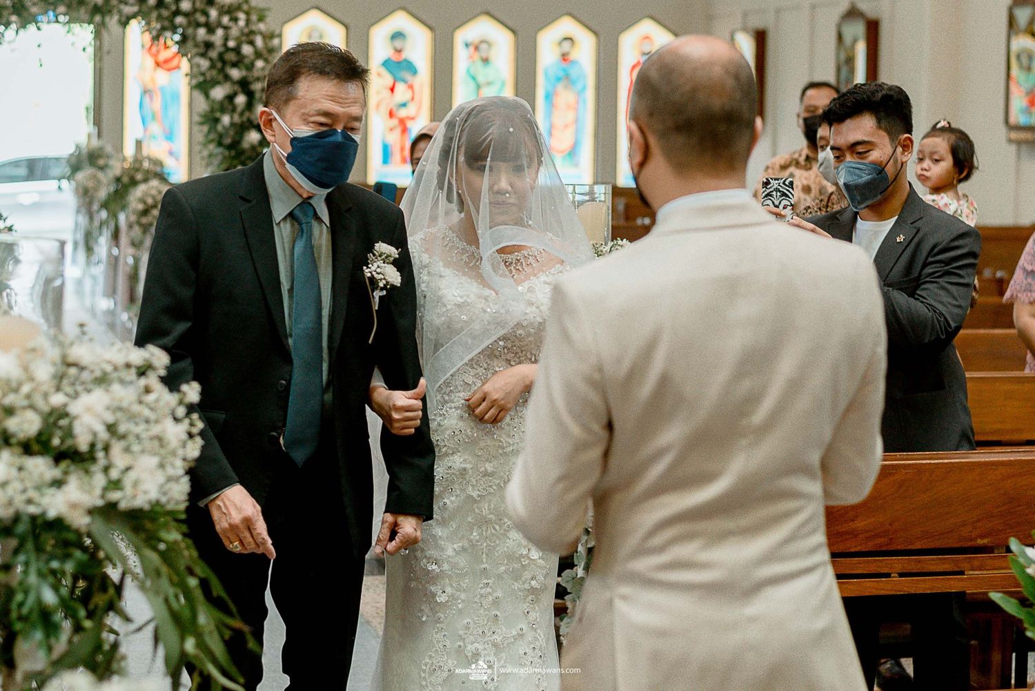 Church wedding ceremony with masked guests during pandemic, featuring stained glass windows and floral decor.
