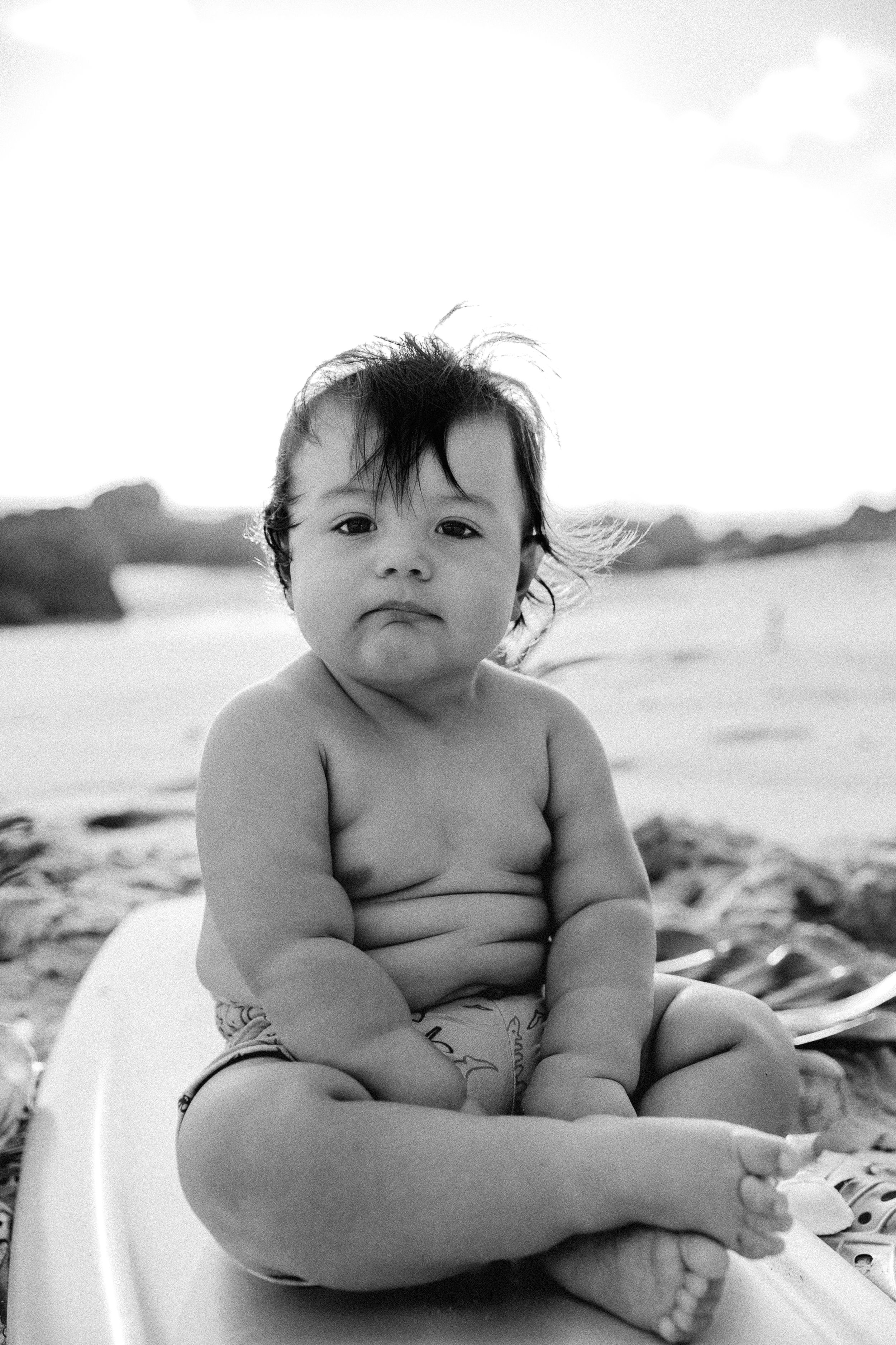 Baby sitting on white surface at beach with rocks in background.
