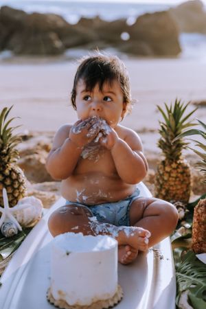 Baby enjoying cake smash on beach with pineapples and ocean backdrop.