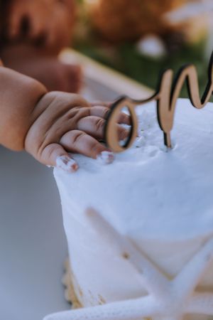 Close-up of small hand touching cake decoration on white frosted surface.
