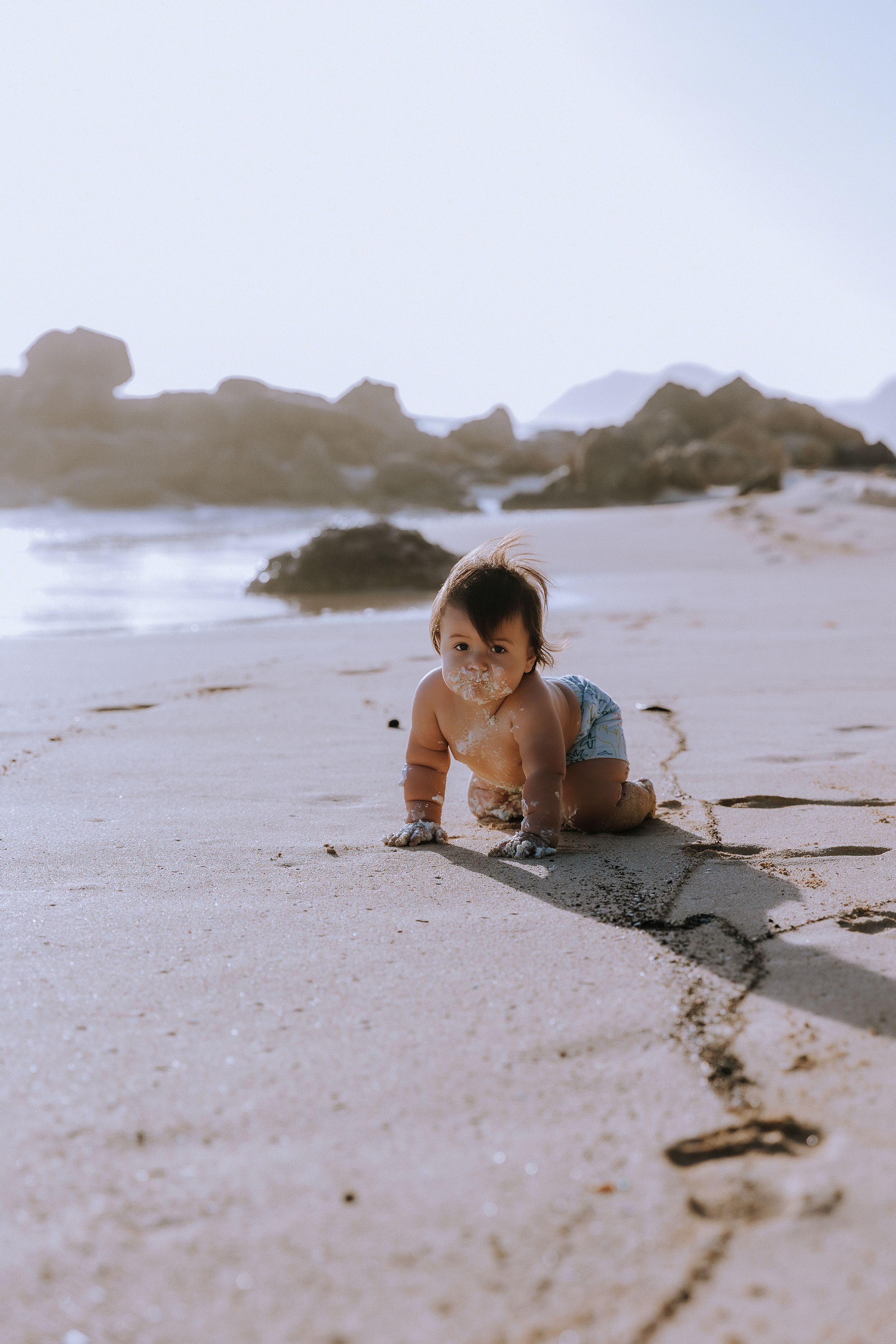 Baby crawling on sandy beach with rocky coastline in background.