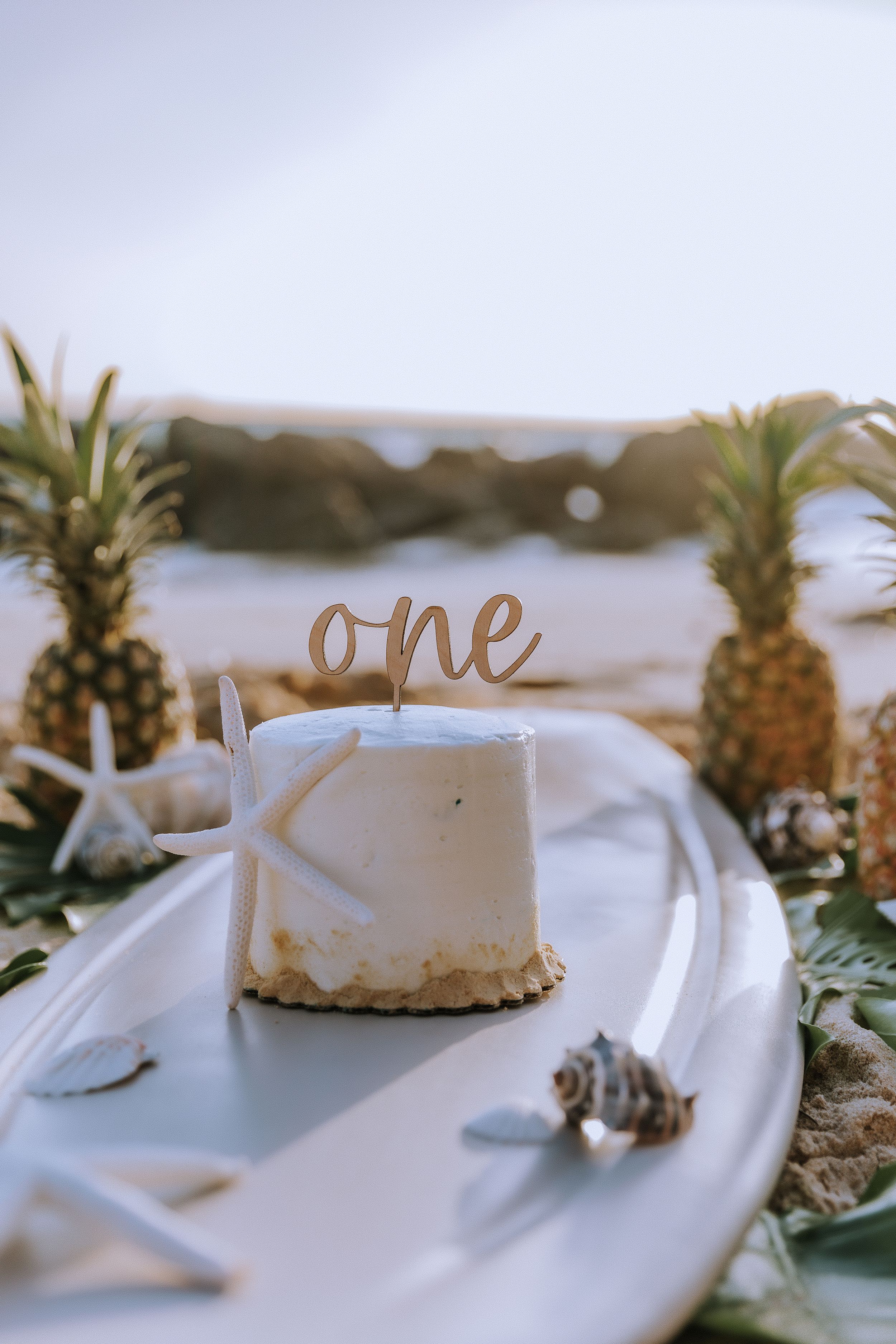 White birthday cake with 'one' topper decorated with pineapples and starfish on beach.