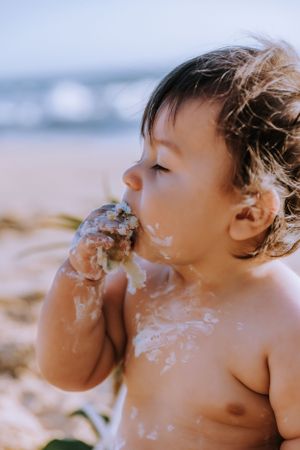 Baby eating cake with messy face against beach background.