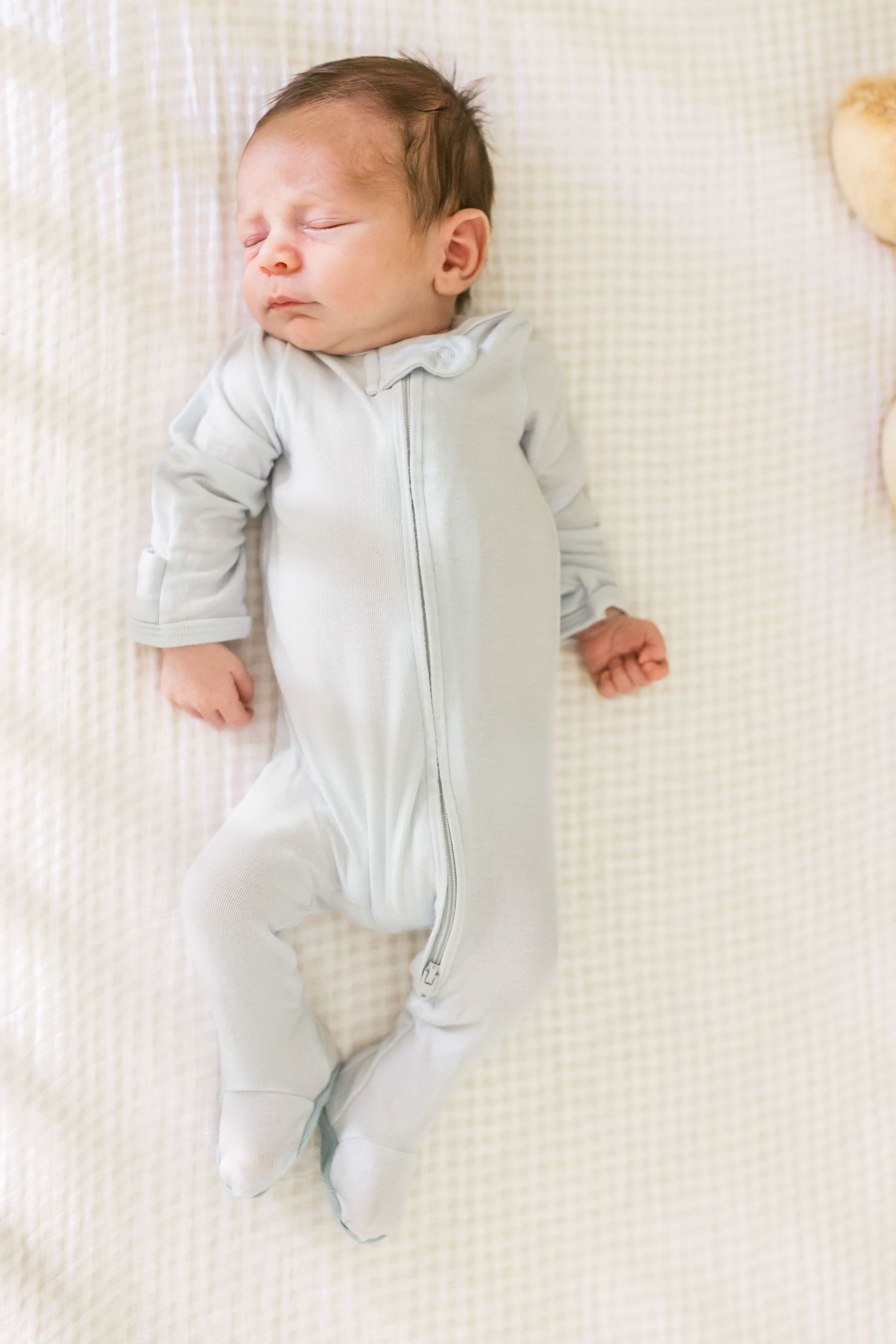 Sleeping baby in light gray onesie lying on white textured bedding.