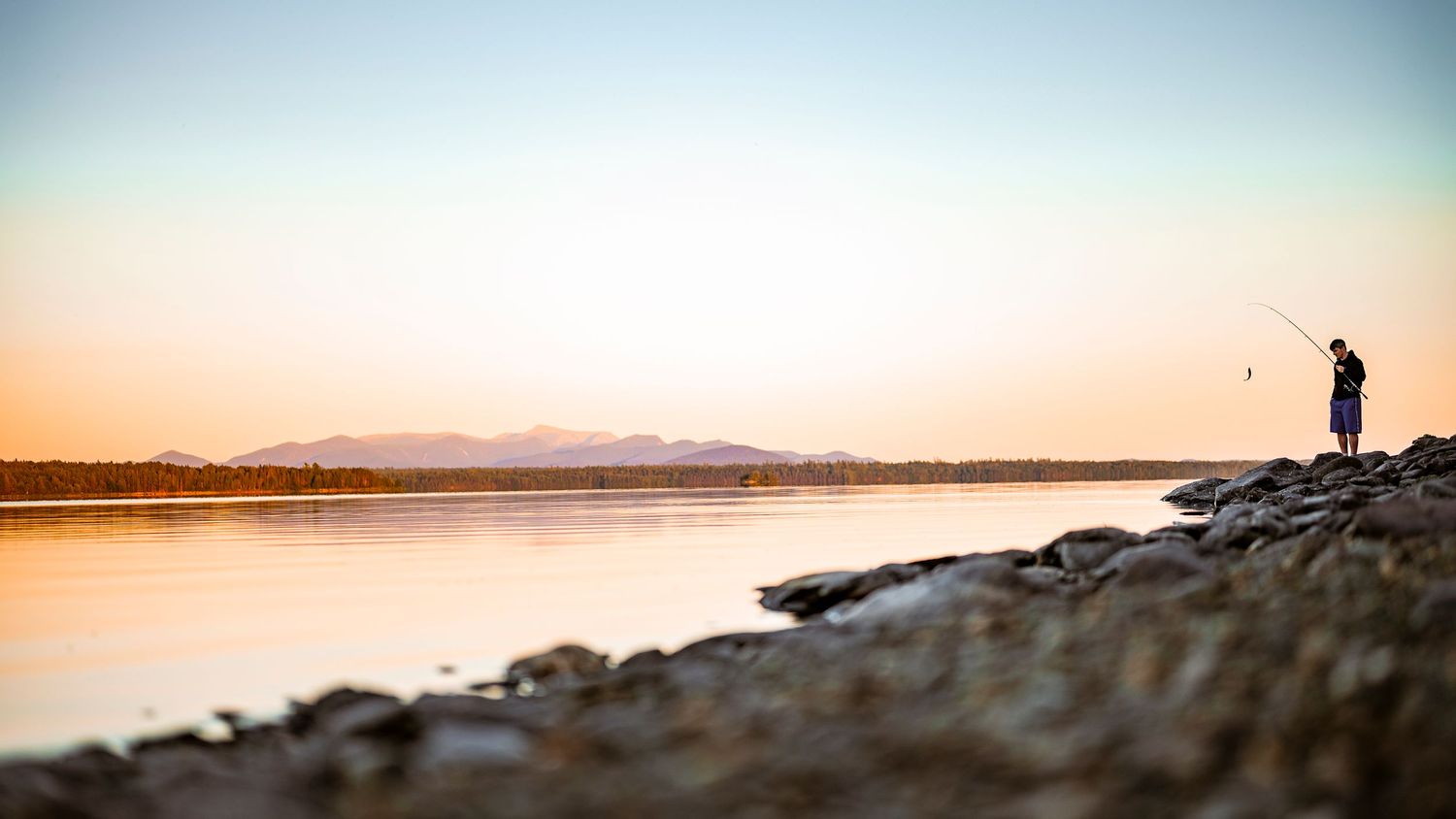 Silhouette of person fishing on rocky shore during sunset with mountains in background.