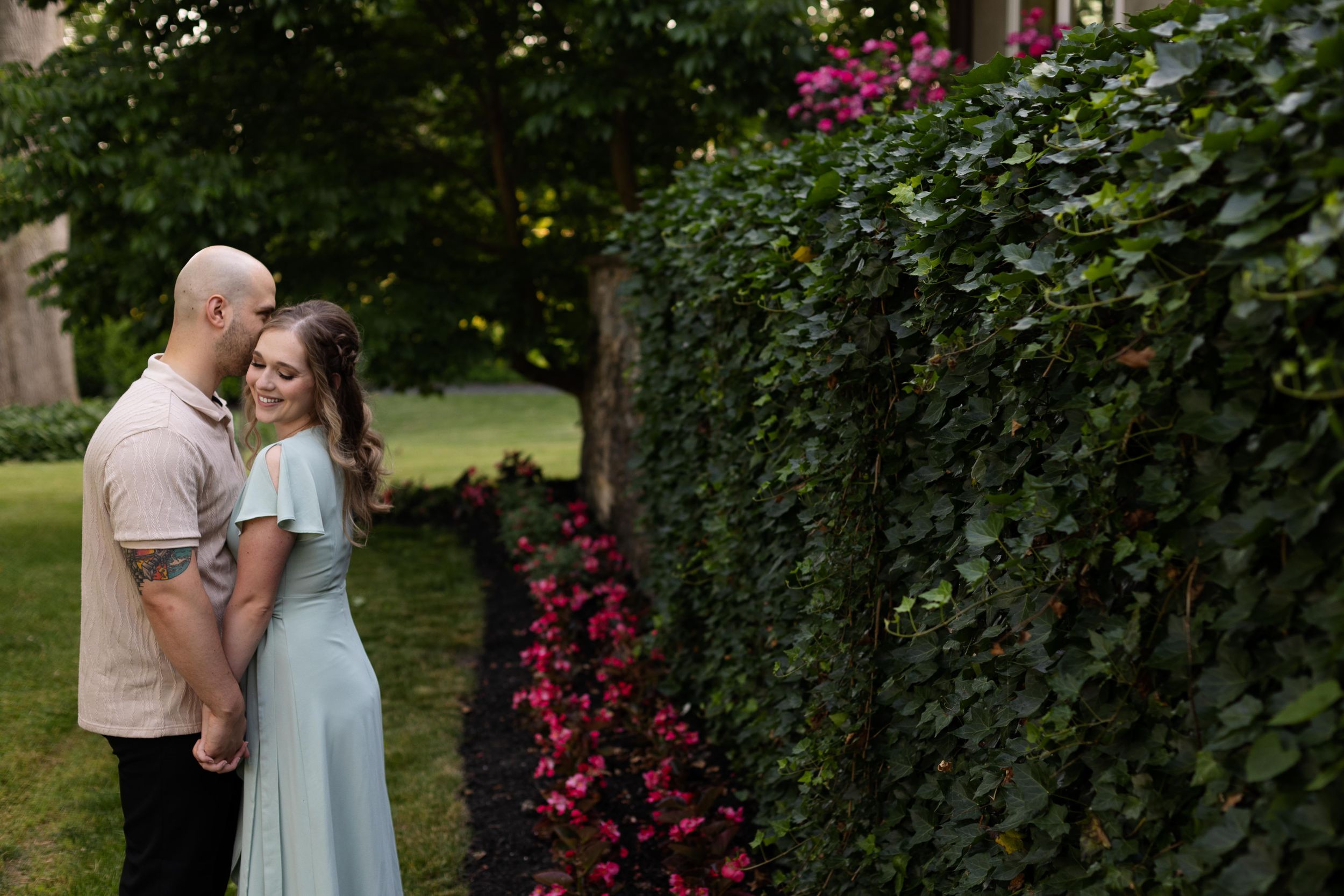 Couple embraces near a landscaped garden with pink flowers and green hedge in summer.