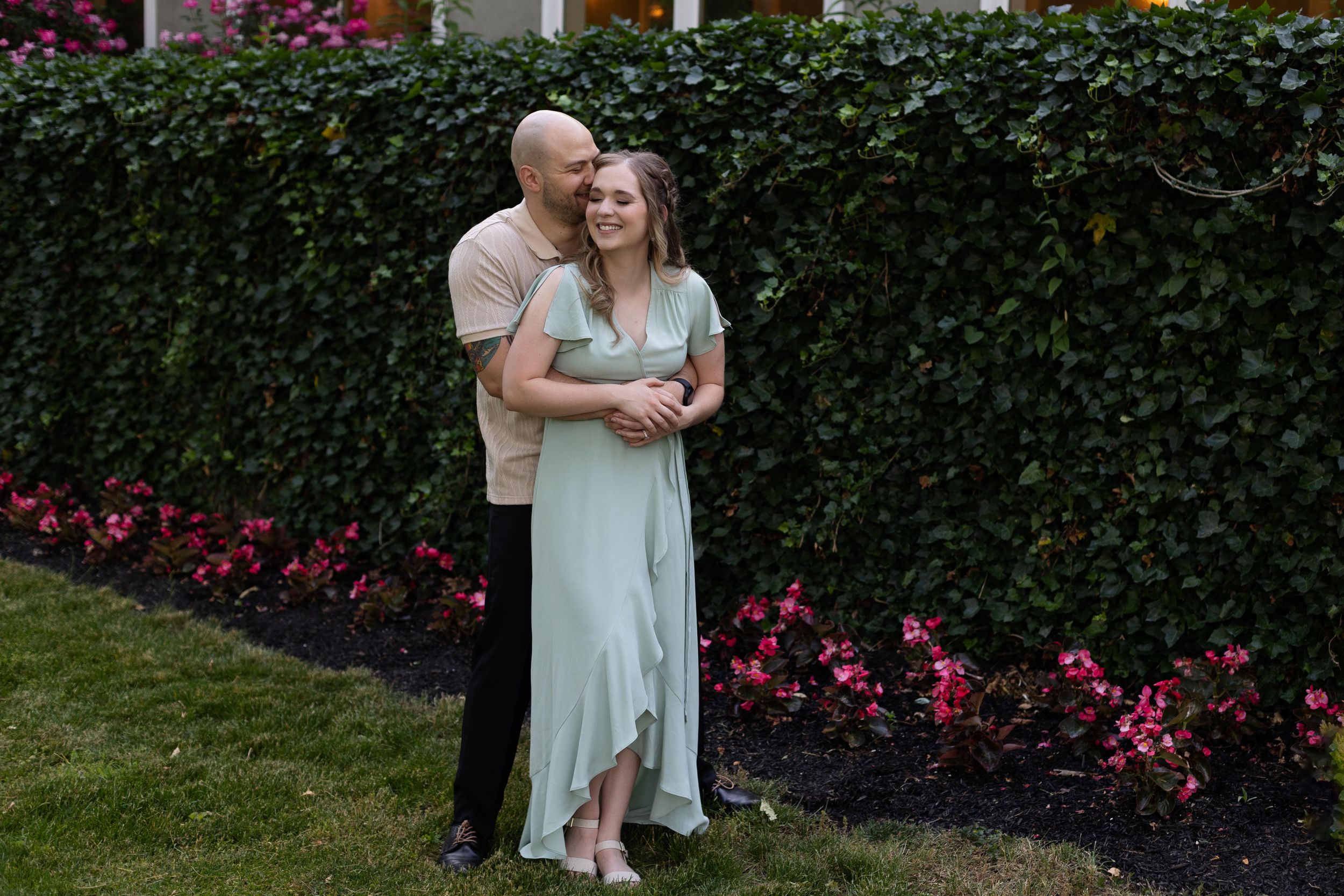 A romantic couple embraces in front of a lush hedge garden with blooming red flowers.