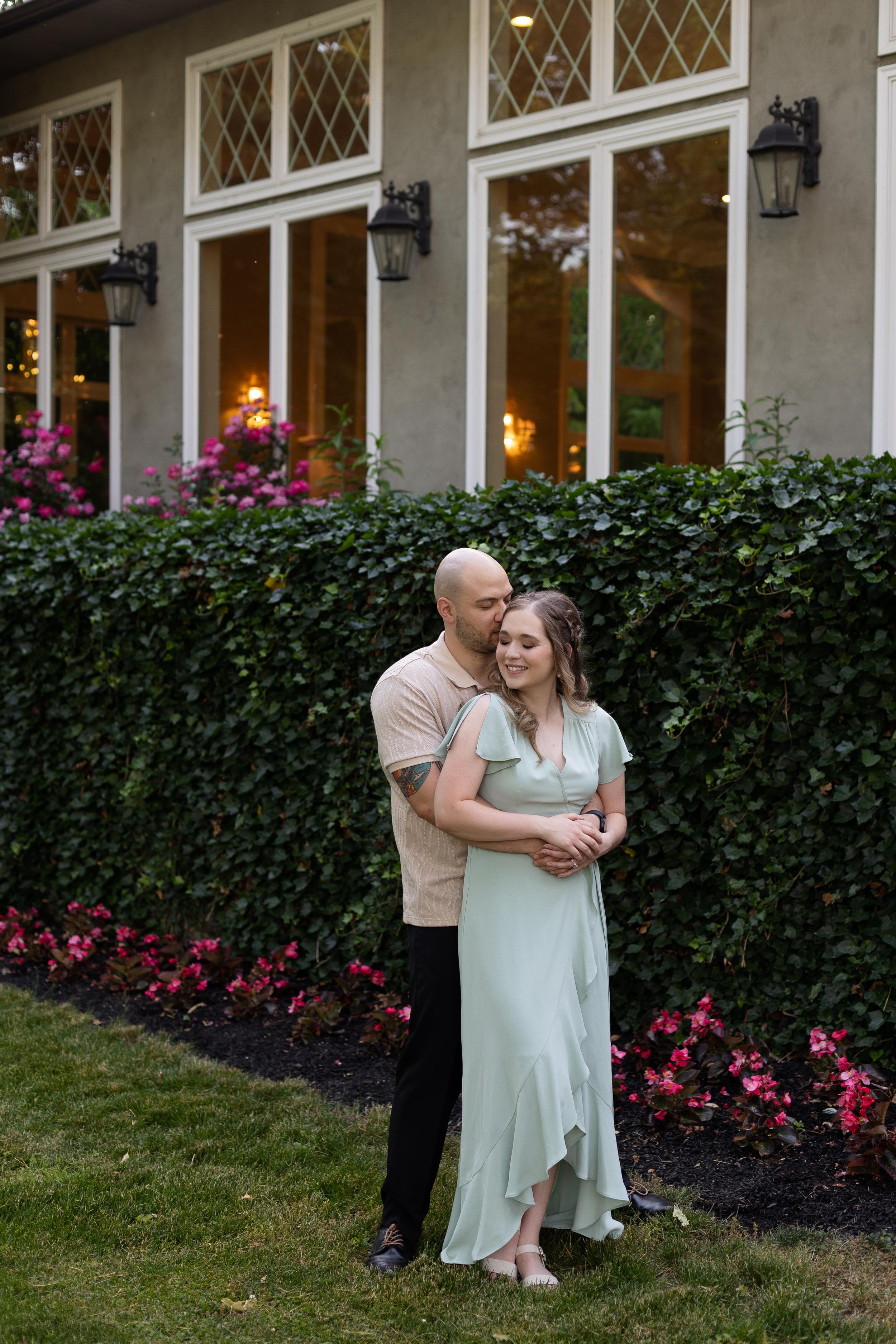 A couple shares an intimate moment against a stately house backdrop with diamond-paned windows.