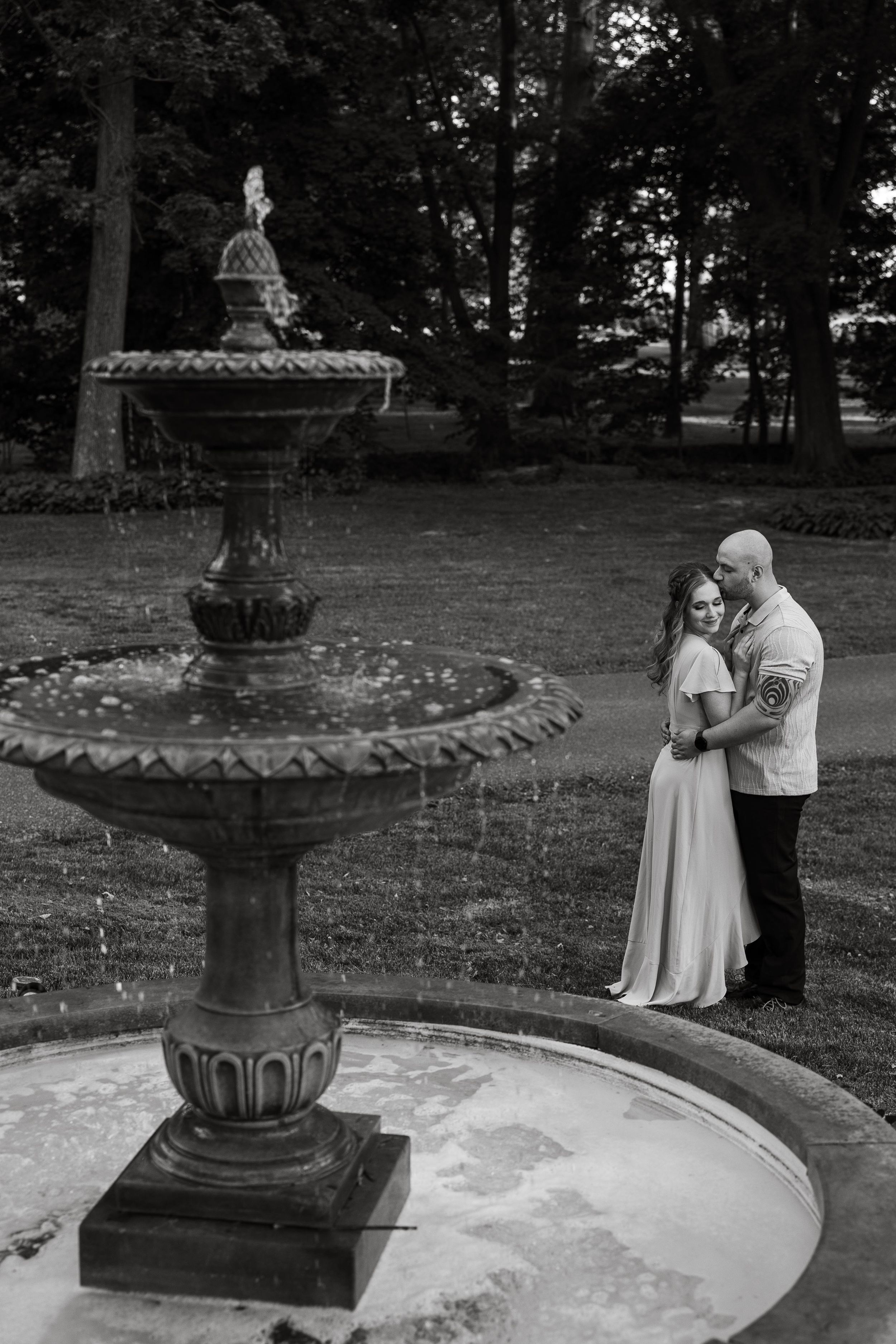 A black and white portrait by an ornate stone fountain in a garden setting.
