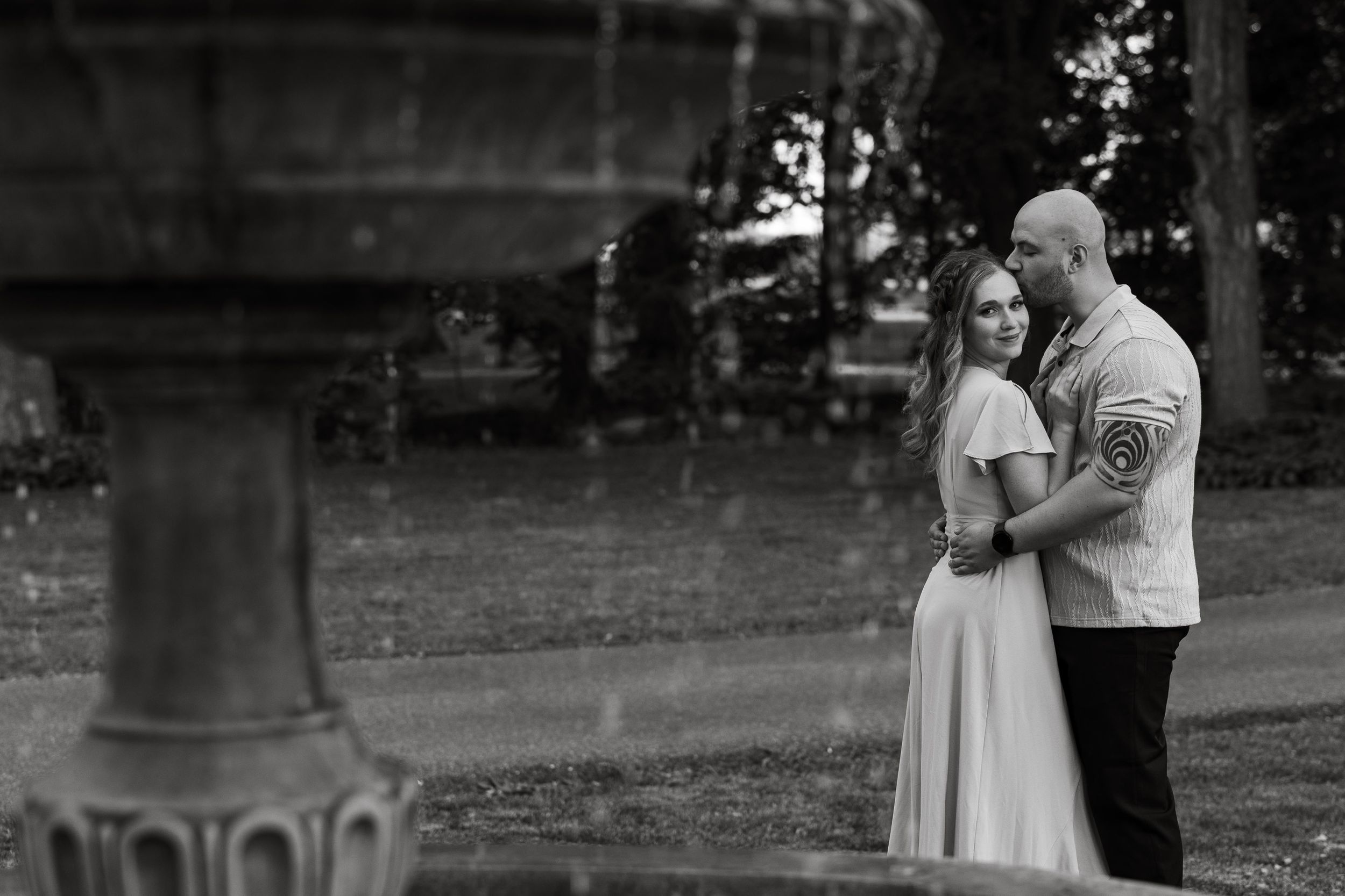 A monochrome portrait of a couple standing near an antique fountain in a park.