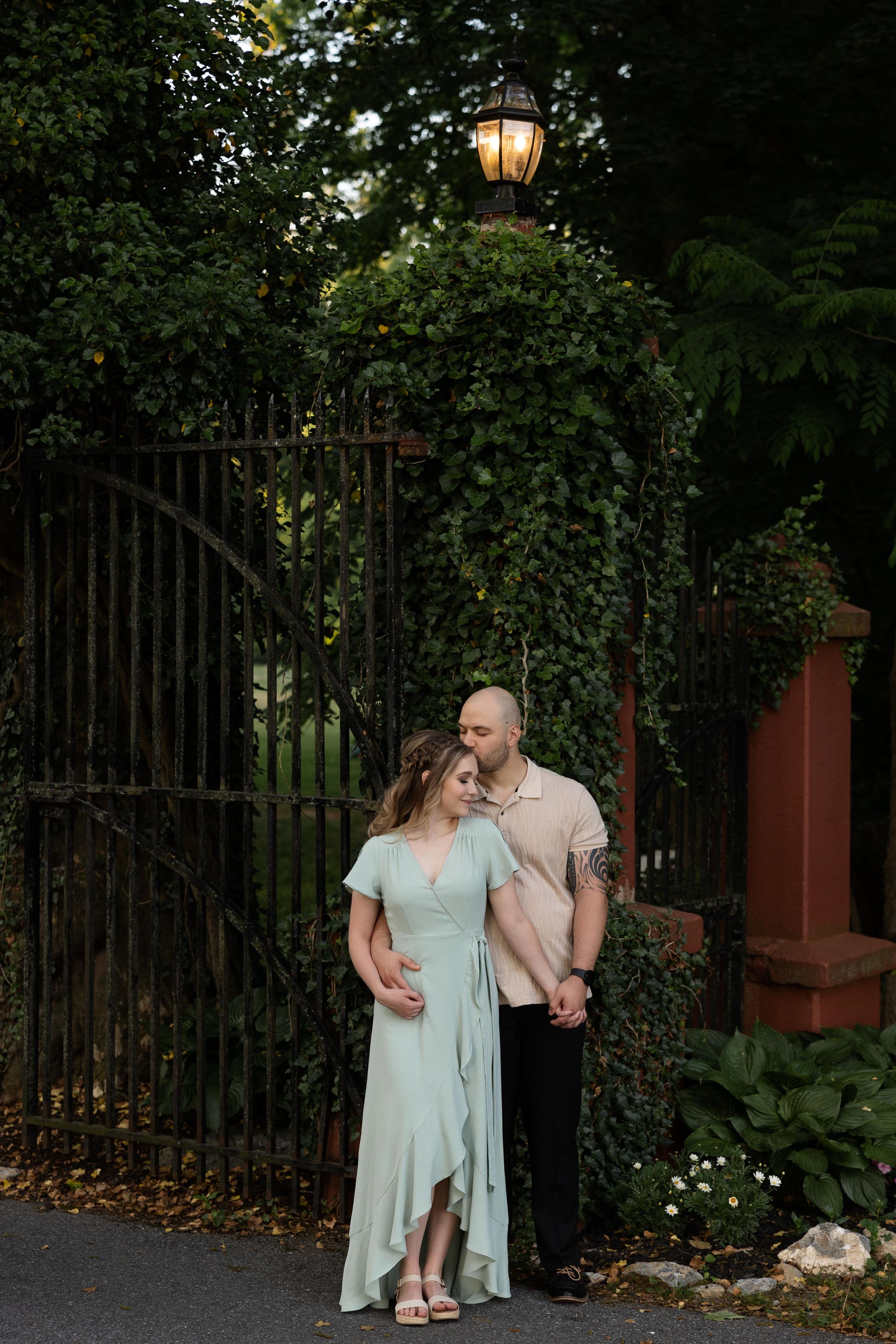 A couple poses beneath a vintage lamp post by an ornate iron gate.