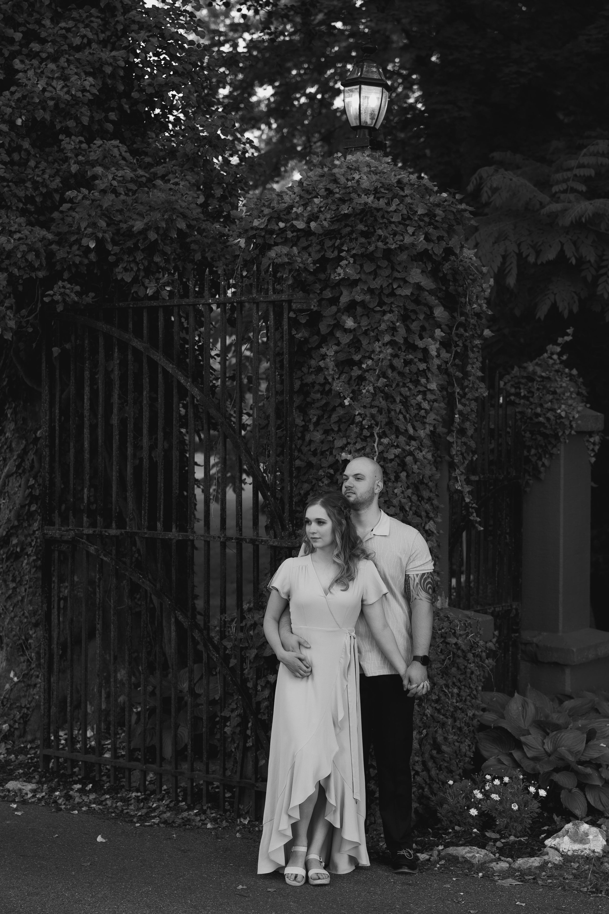 A romantic black and white photo of a couple standing by an ornate iron gate with climbing vines.