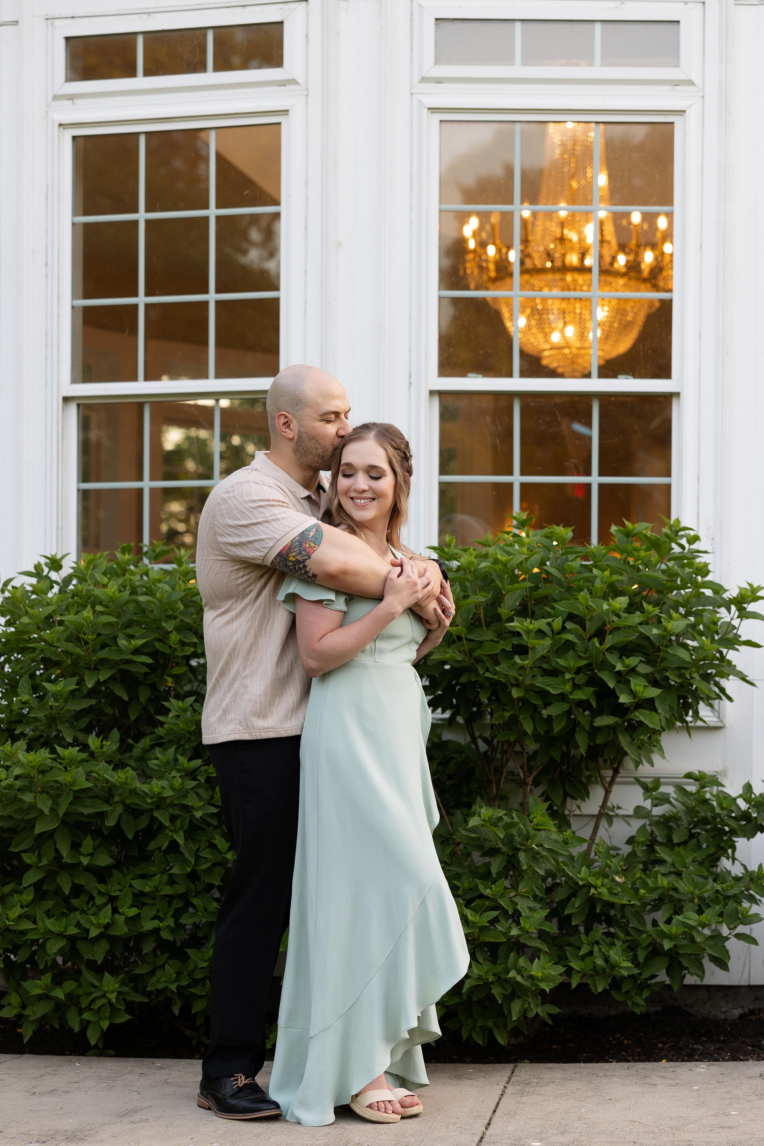 Couple embracing outside white windows with warm chandelier glow visible inside.