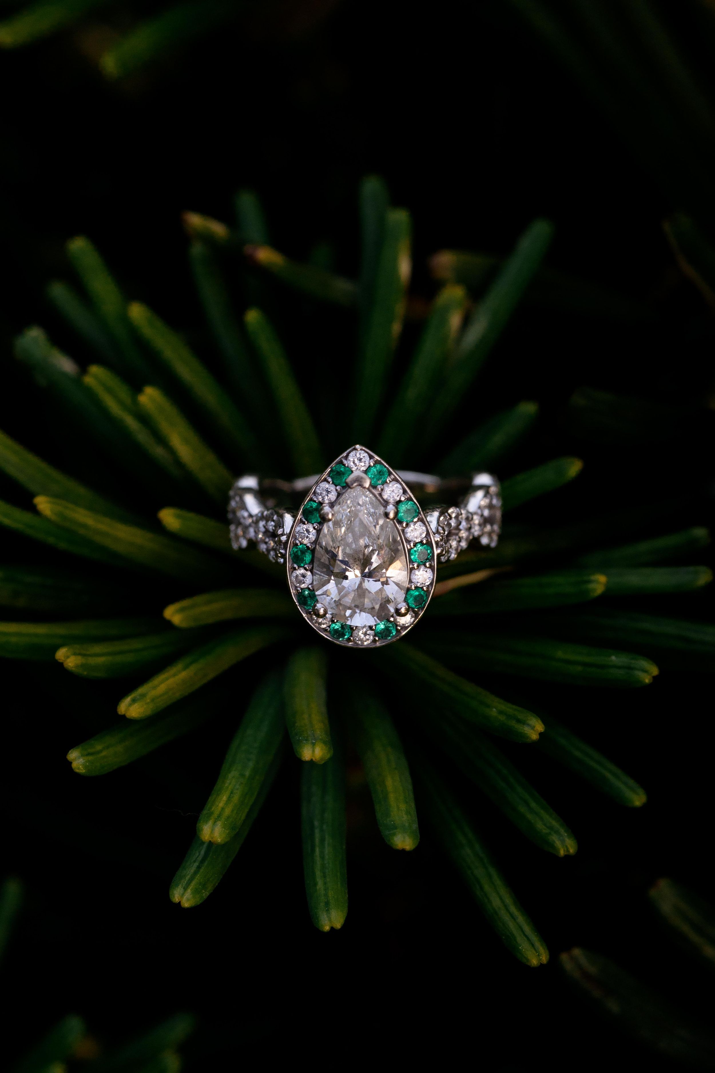 Vintage-style engagement ring with emerald accents photographed on dark green foliage.
