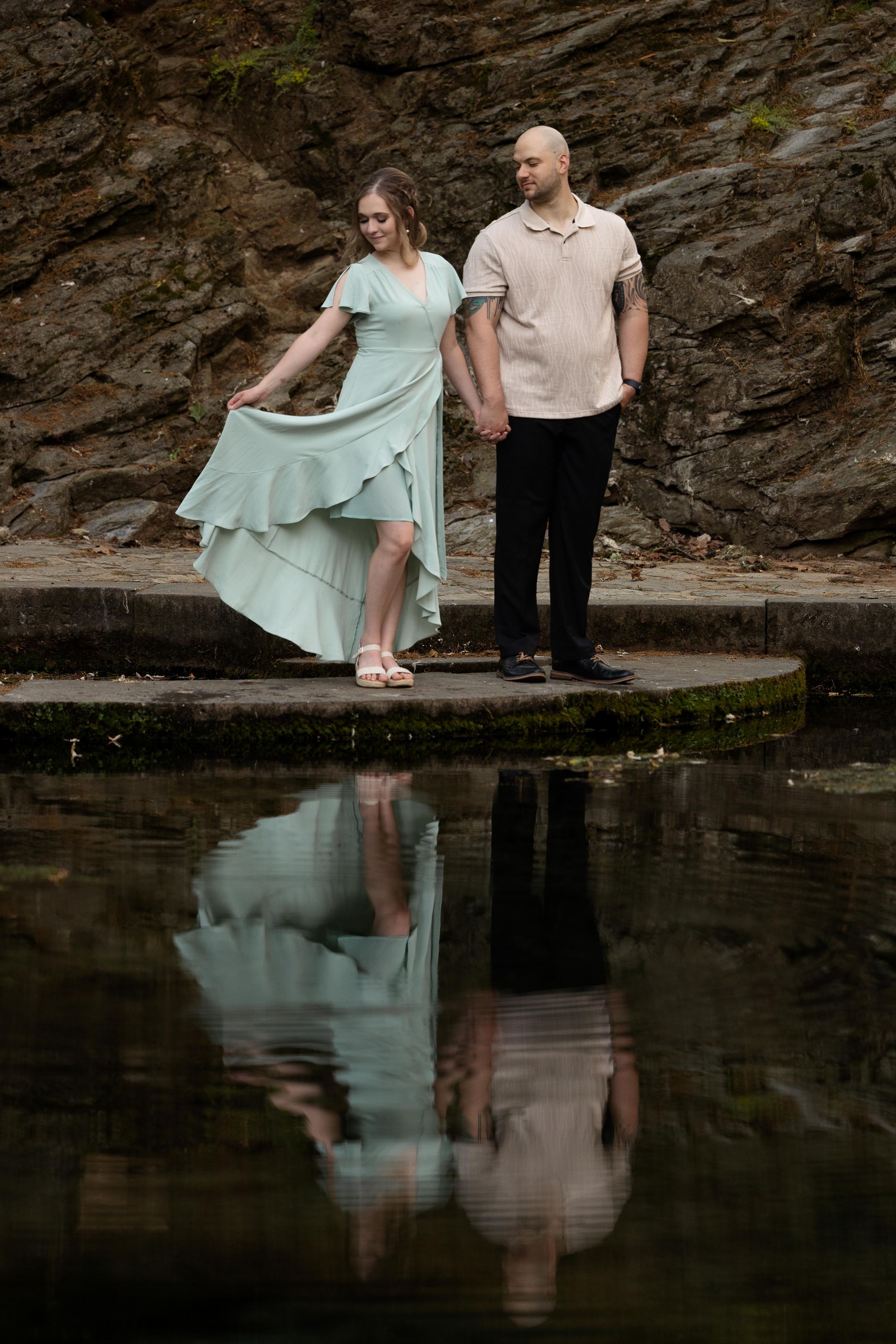 A romantic couple in mint and beige stands by a pond with their reflection visible in the still water.
