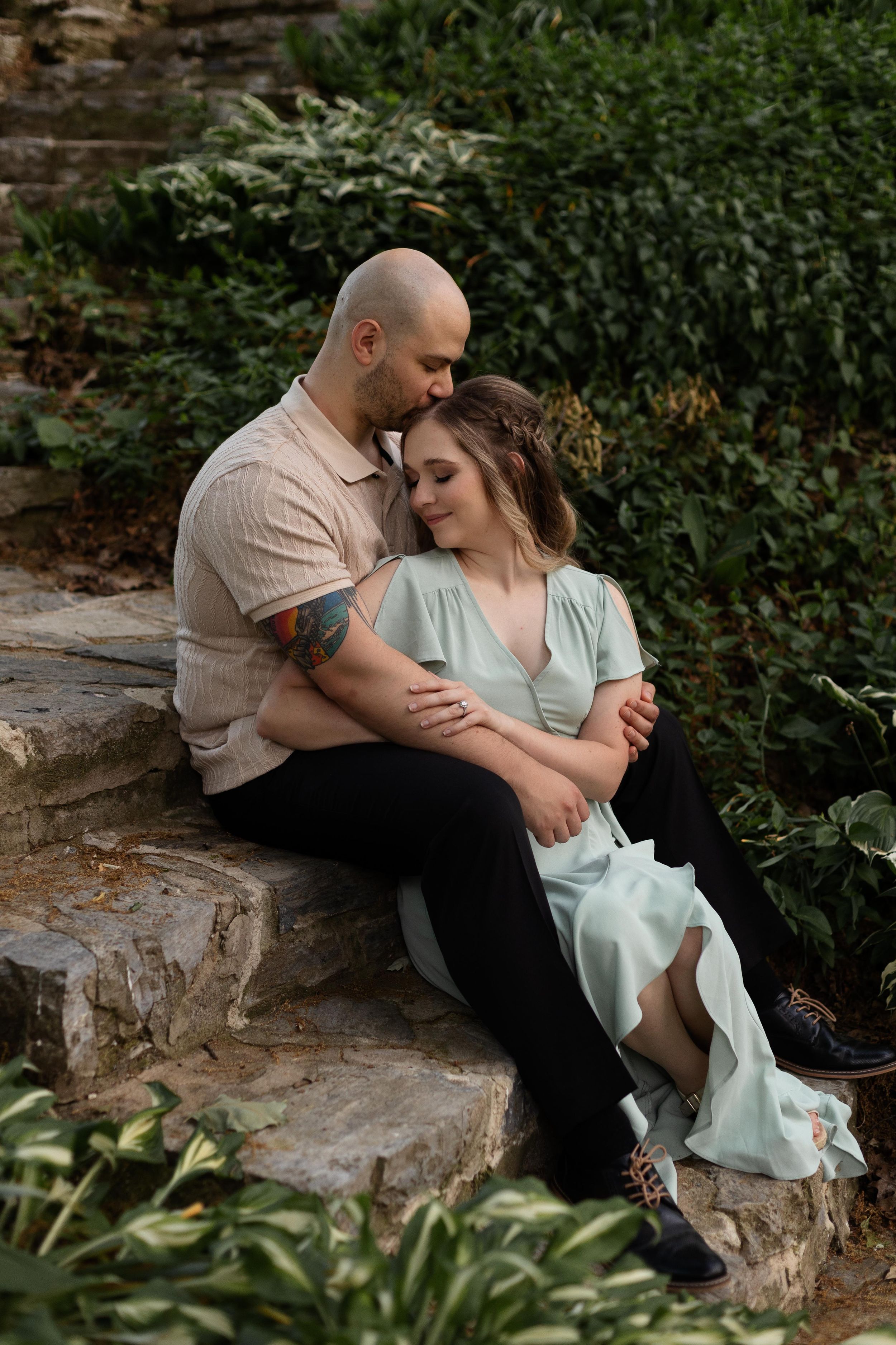 A couple in pastel clothing shares a tender moment while sitting on stone steps in a garden setting.