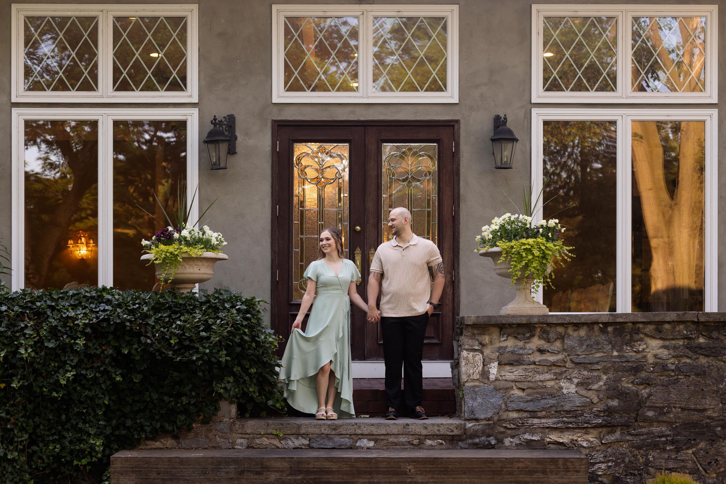 Couple stands on stone steps in front of a grand house with large windows and lanterns.