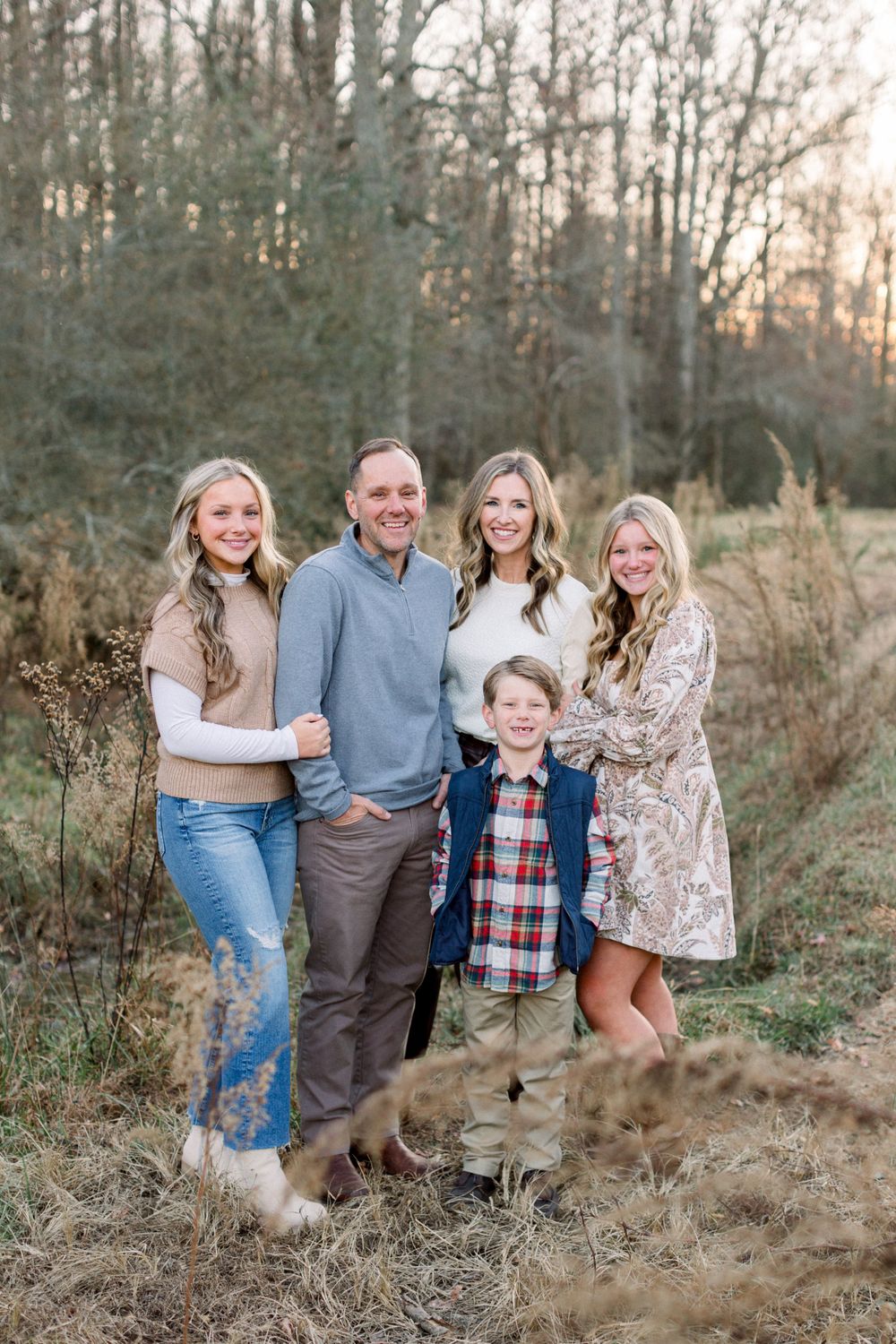 A family of five poses together smiling in a golden field at sunset during autumn season.