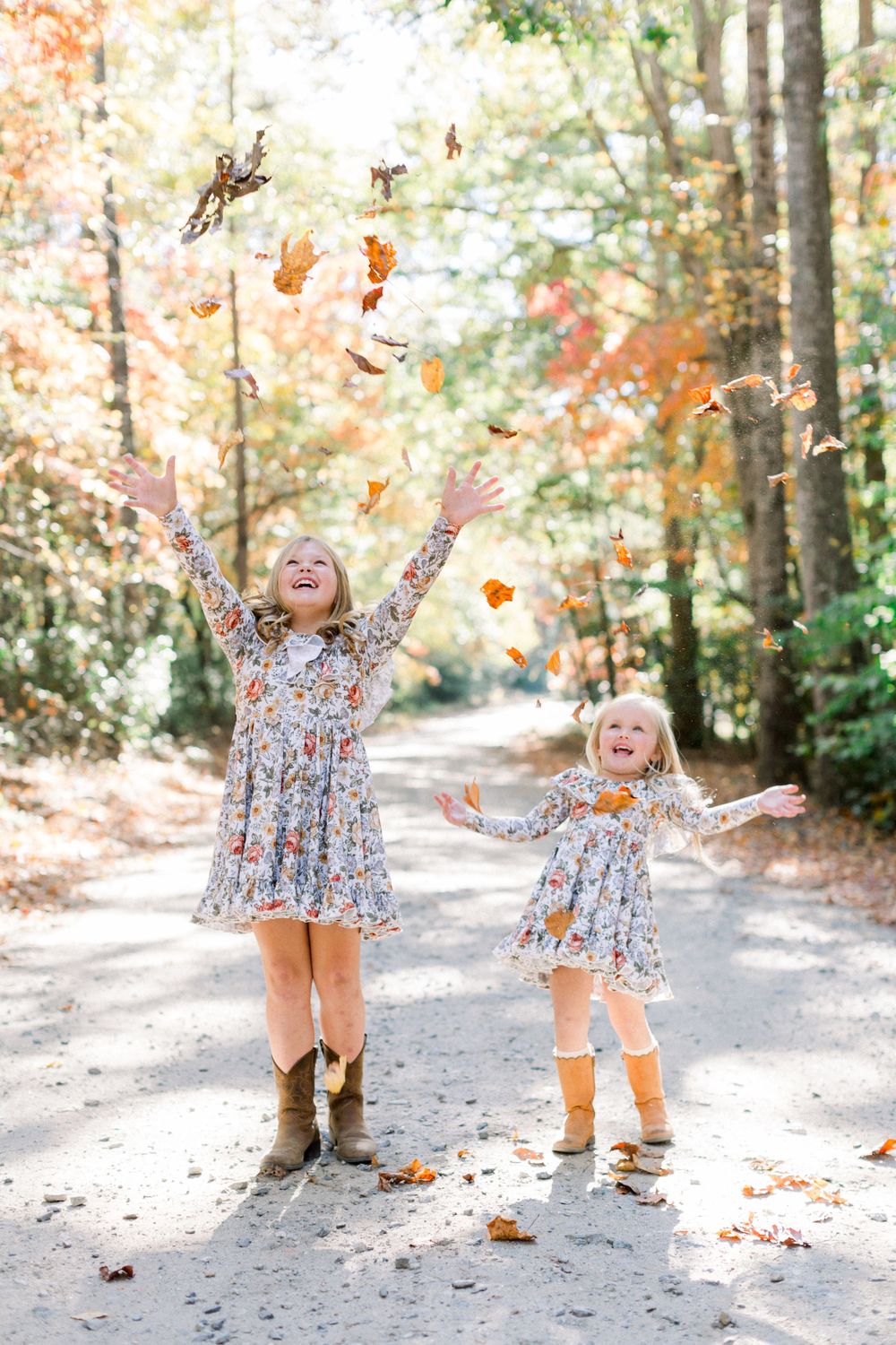 Two young girls in floral dresses joyfully toss autumn leaves on a sunlit forest path.
