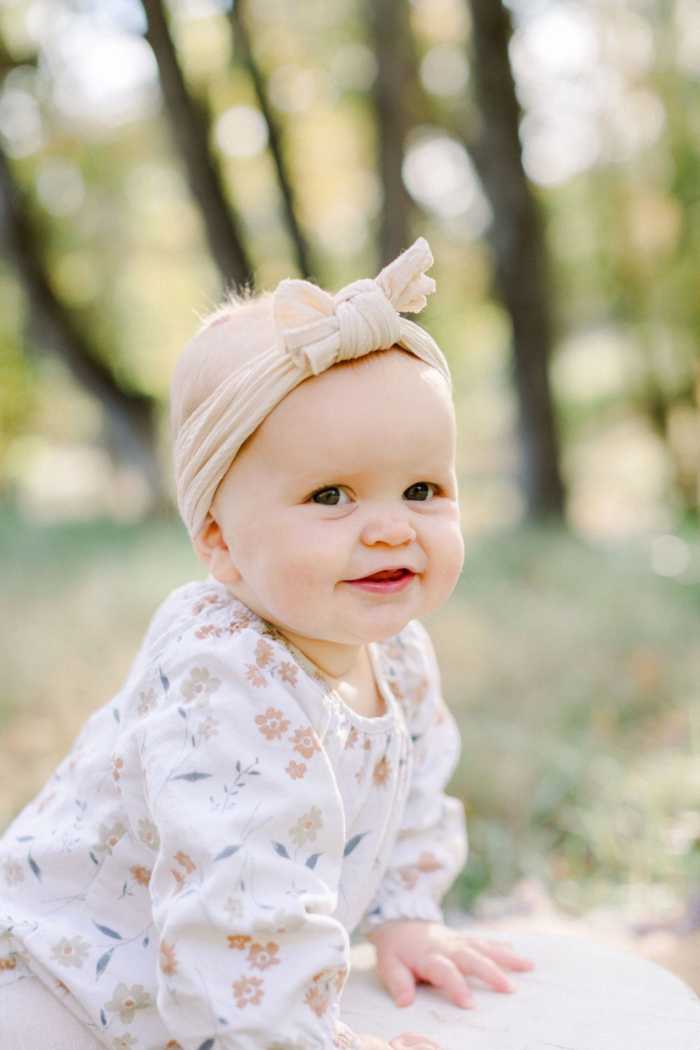 Adorable baby girl in a floral outfit and beige bow headband smiles outdoors in a sunlit park setting.