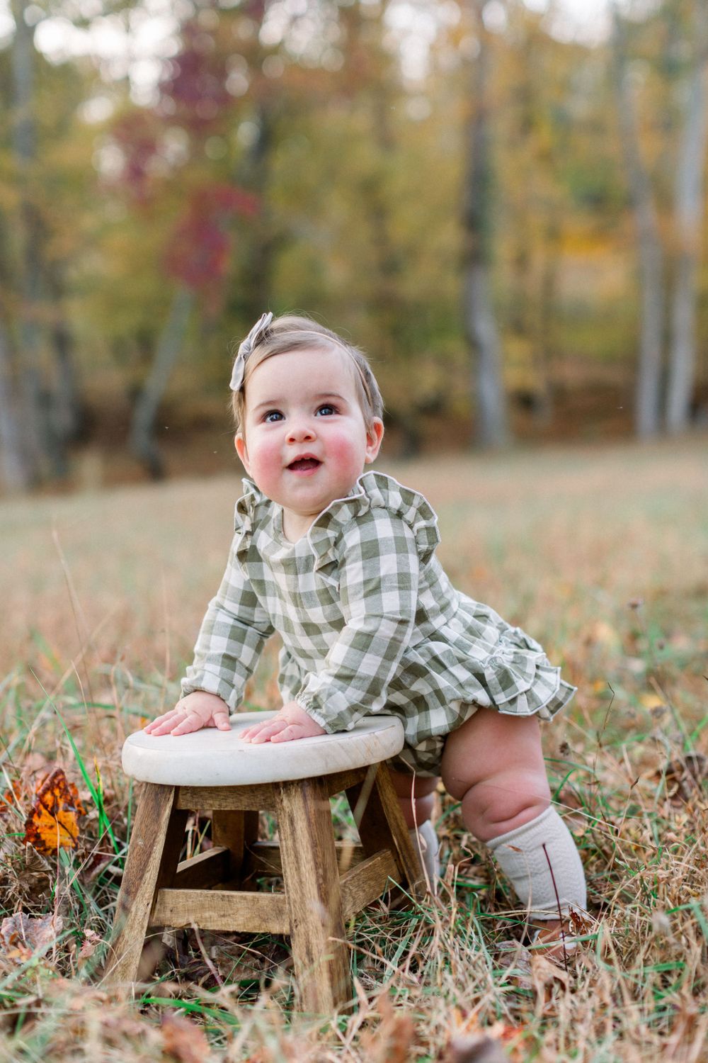 An adorable baby in a green plaid dress stands beside a small wooden stool in an autumn park.