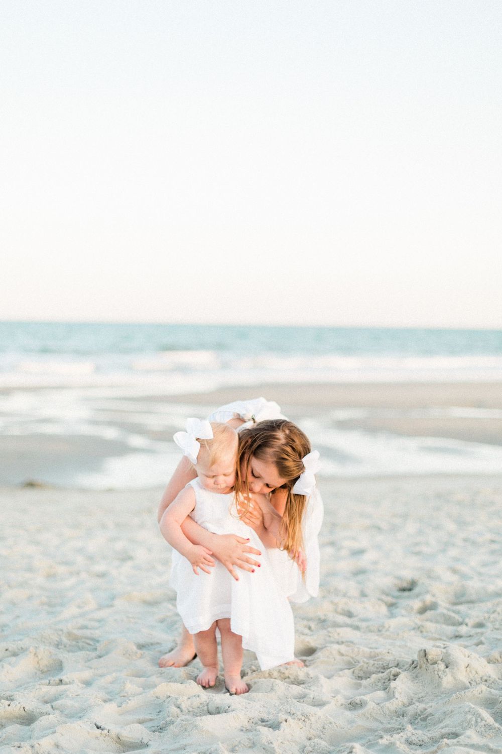 A tender moment as a mother in a white dress hugs her toddler daughter on a serene beach at sunset.