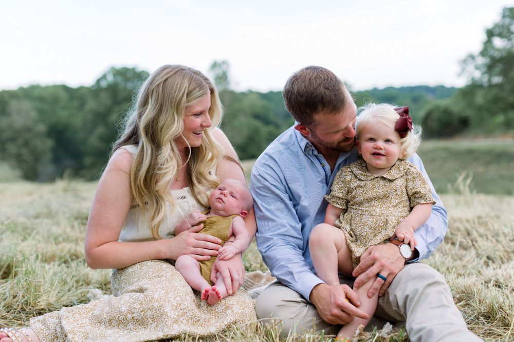Happy young family sits in a green meadow, parents smiling at their newborn and toddler daughter.
