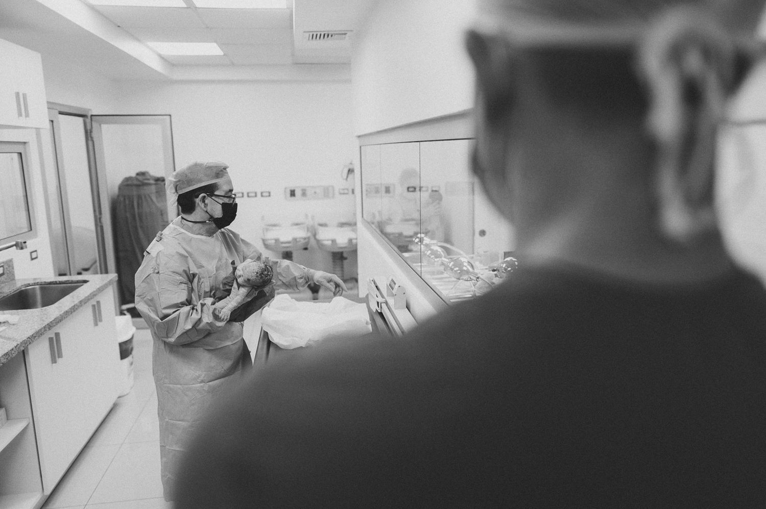 Healthcare workers in protective gear stand in hospital nursery with rows of newborns.