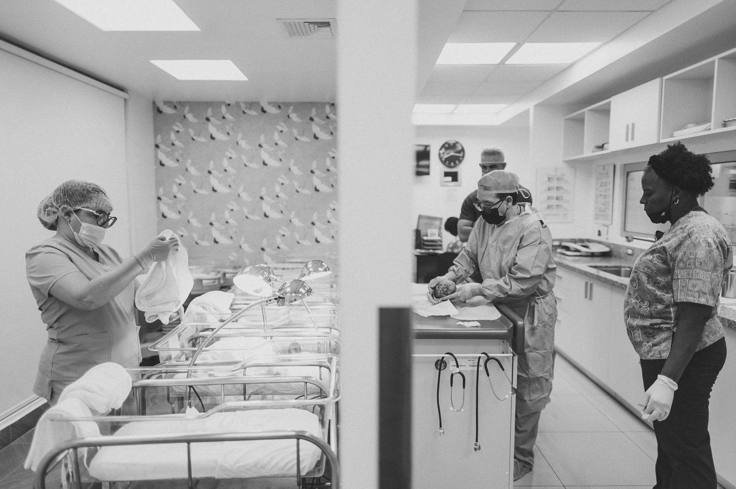 Medical staff in masks working in hospital nursery with multiple newborn bassinets.