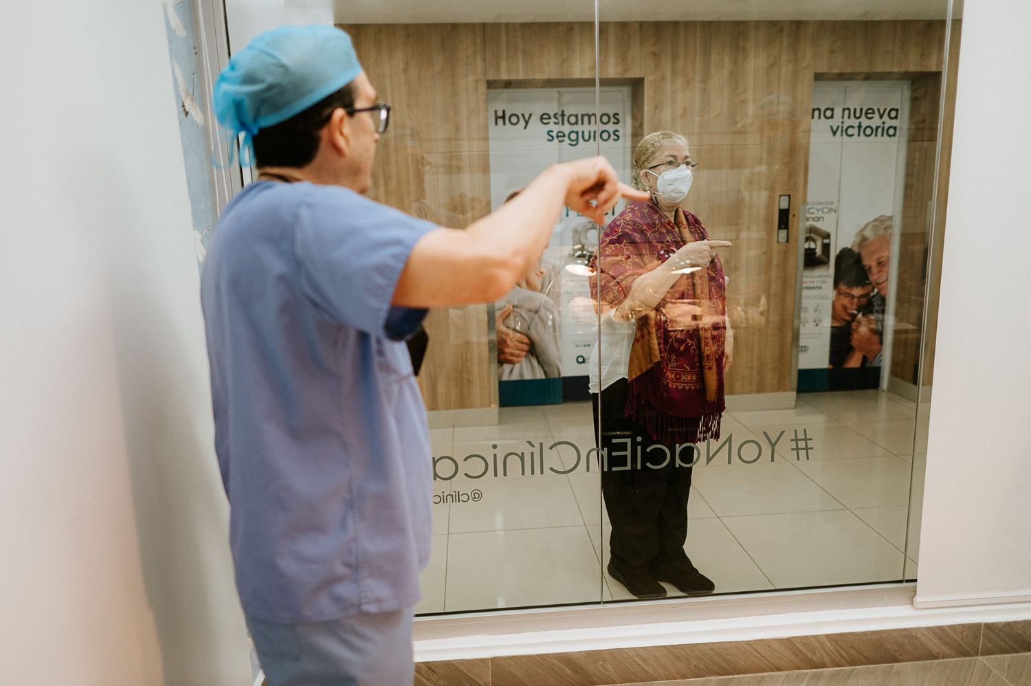 Healthcare worker in blue scrubs communicates through glass window in medical setting.