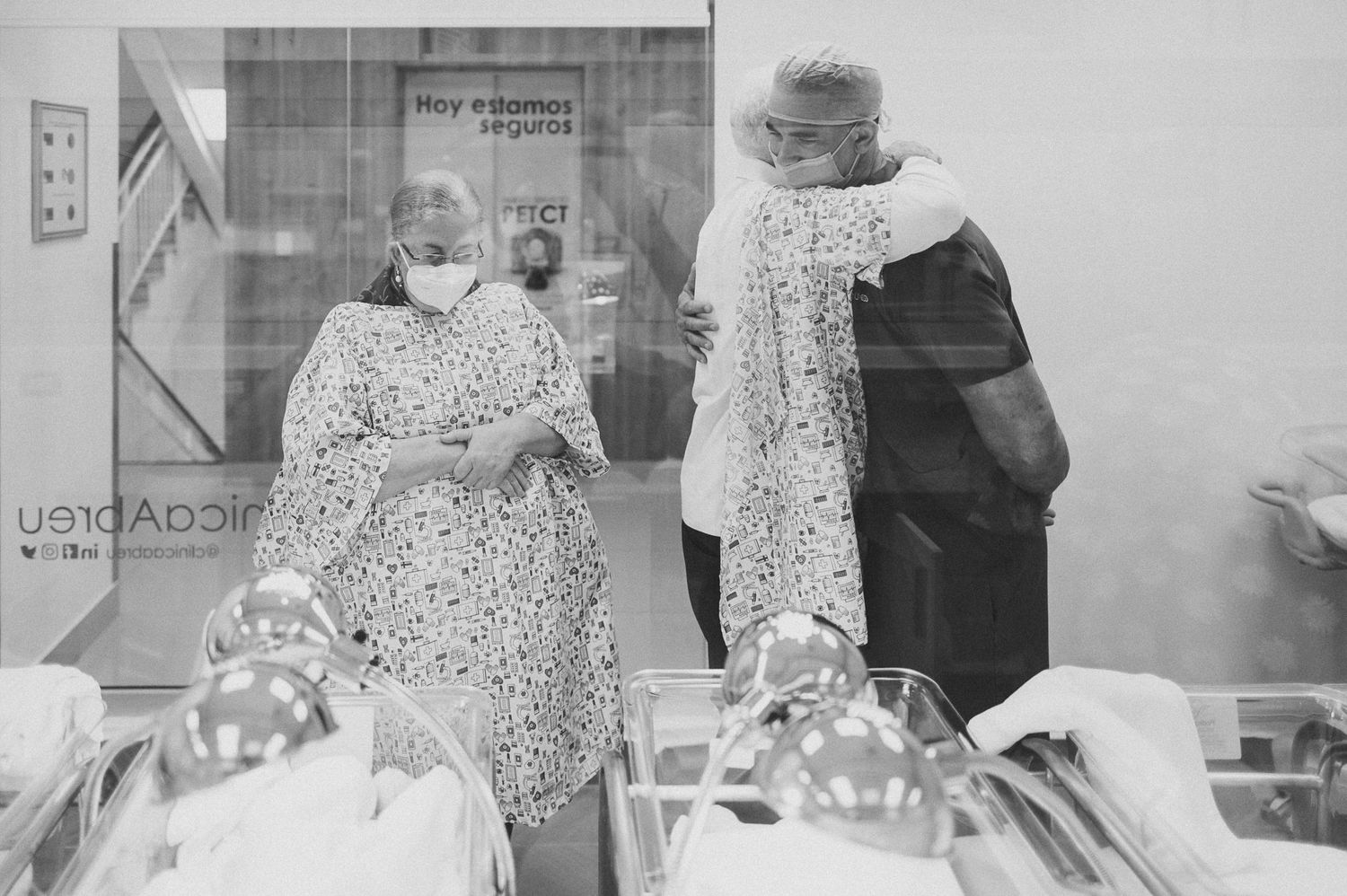 Two people wearing patterned garments observe newborn infants through a hospital nursery window.