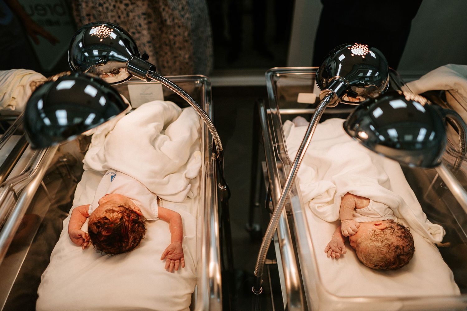 Two newborn babies rest under warming lamps in hospital bassinets.