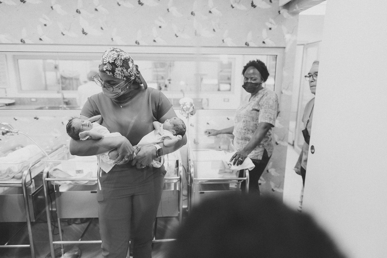 Person holding a newborn baby in a hospital nursery with bassinets visible.