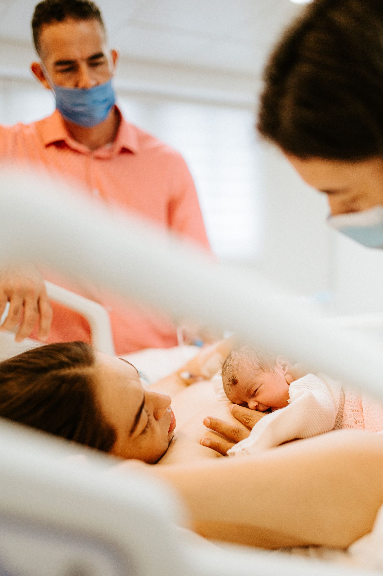 Healthcare worker in a pink shirt wearing a mask watches over a newborn baby in a hospital room.