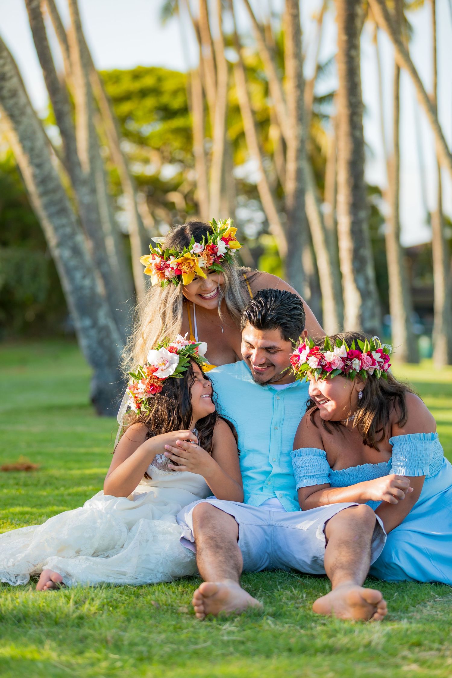 Family wearing flower crowns relaxing together on grass under palm trees in Hawaii.