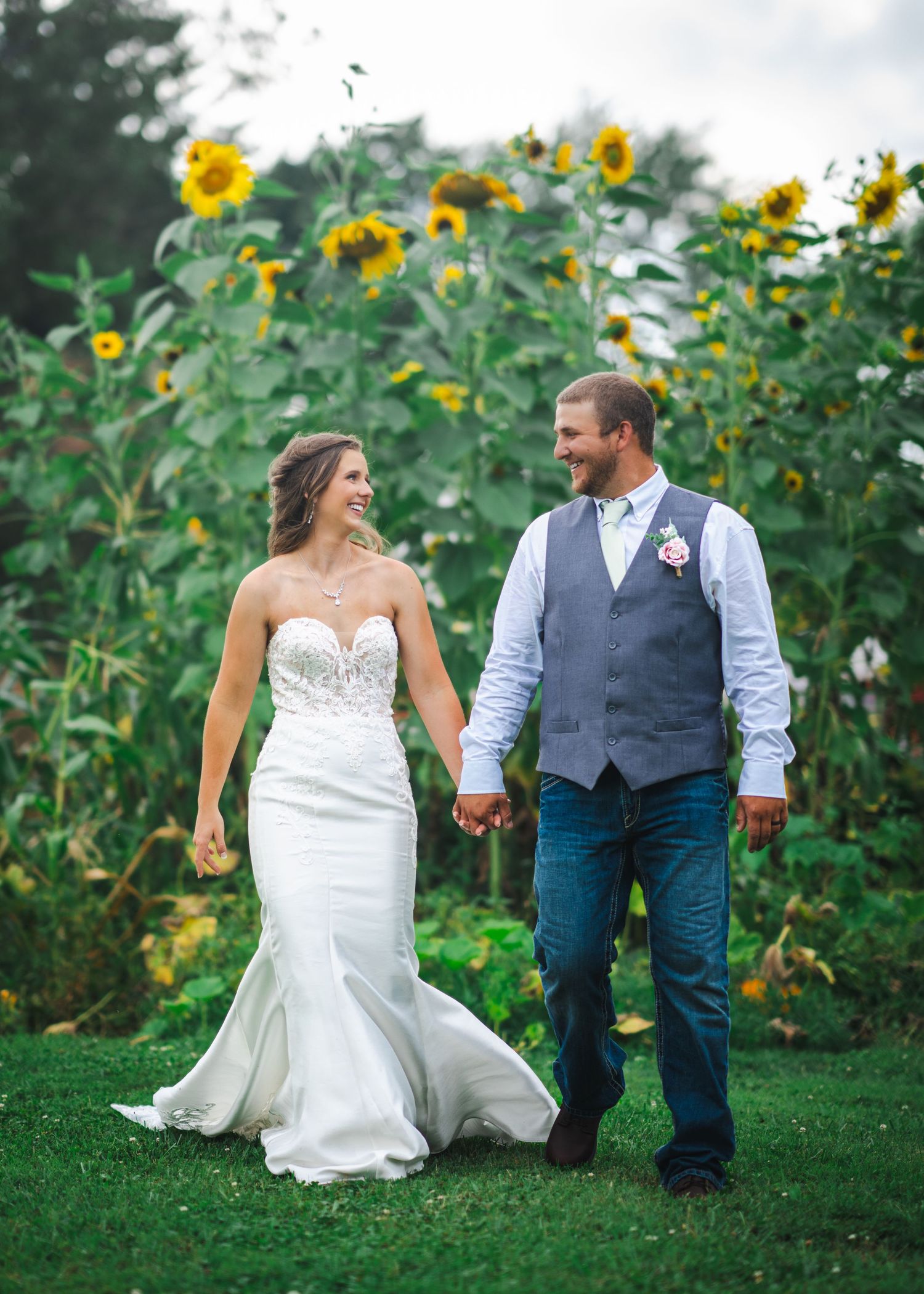 Bride and groom walk hand in hand through garden with towering sunflowers in background.