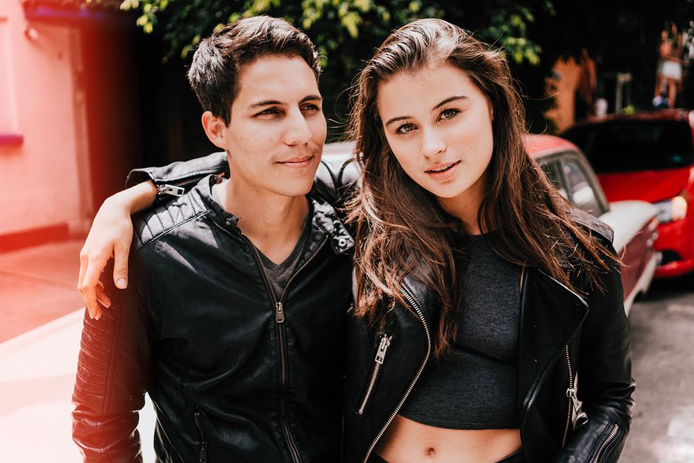 A casual couple in leather jackets posing against a coral-colored wall.